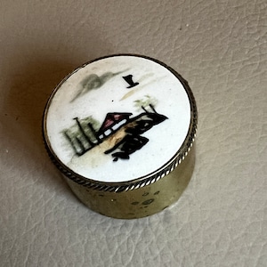 Puede incluir: Una pequeña caja redonda de latón con una tapa de porcelana blanca. La tapa tiene una escena pintada a mano de una casa con un techo rojo, árboles y una nube. El número "1" está pintado en la tapa.