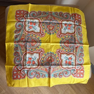 Può includere: Una sciarpa quadrata gialla con un elaborato motivo floreale e paisley. La sciarpa presenta uno sfondo giallo brillante con motivi ornati in rosso, verde acqua e bianco. Il design include bouquet floreali ed elementi paisley a spirale.
