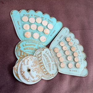 May include: A collection of vintage button cards. The cards are light blue with gold text, reading "6 Assorted Sizes Superior Quality Buttons". The buttons are off-white and arranged on the cards, made in England.