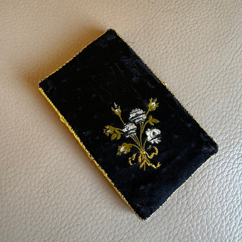 May include: A black velvet pouch with a gold trim and a white floral embroidery design.