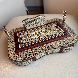 Pode incluir: Um conjunto de secret&aacute;ria em madeira ornamentada com um padr&atilde;o de mosaico floral em tons de castanho, dourado e preto. O conjunto inclui um porta-canetas, um porta-blocos de notas e uma superf&iacute;cie de escrita central com uma incrusta&ccedil;&atilde;o decorativa. Inclui duas canetas douradas.