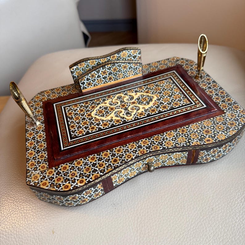 Pode incluir: Um conjunto de secret&aacute;ria em madeira ornamentada com um padr&atilde;o de mosaico floral em tons de castanho, dourado e preto. O conjunto inclui um porta-canetas, um porta-blocos de notas e uma superf&iacute;cie de escrita central com uma incrusta&ccedil;&atilde;o decorativa. Inclui duas canetas douradas.