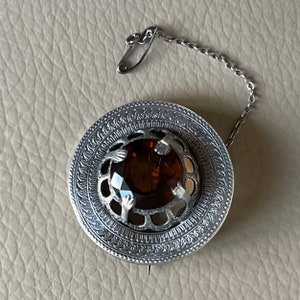 May include: A silver brooch featuring a brown gemstone set within a Celtic knot design. The brooch has a safety pin fastening and a chain attached.