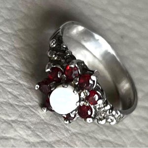May include: A silver ring featuring a cluster of red gemstones and a central white gemstone. The ring has a textured design around the gemstones and a smooth, polished band. A piece of jewelry.