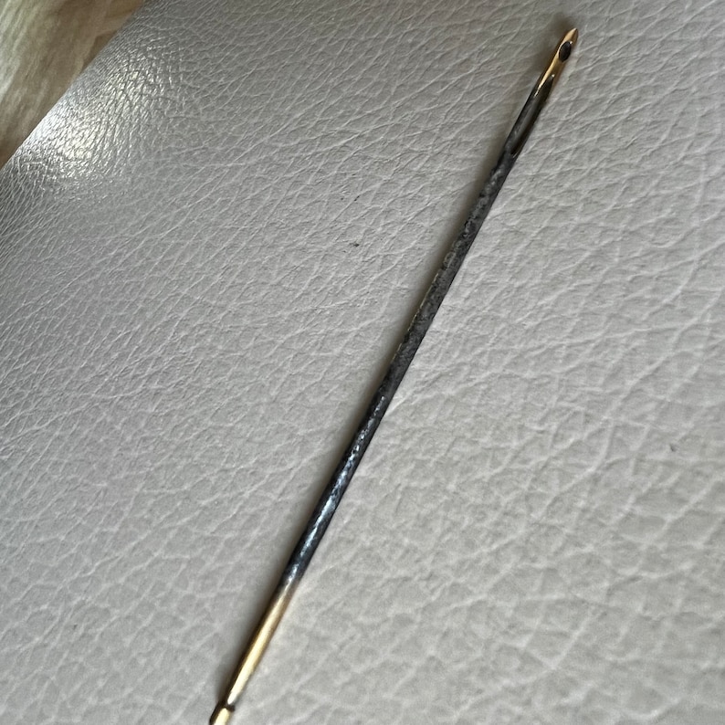 Pode incluir: Uma agulha longa e fina de metal com uma ponta dourada. A agulha est&aacute; ligeiramente dobrada e tem uma p&aacute;tina cinza escura.