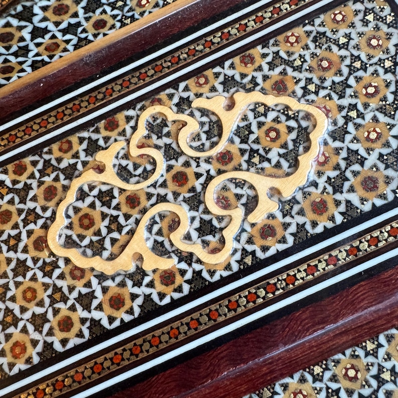 Pode incluir: Close-up de uma superf&iacute;cie de madeira decorativa com intrincados padr&otilde;es embutidos. O design apresenta formas geom&eacute;tricas em preto, dourado e branco, com uma pe&ccedil;a de destaque clara, ornamentada e em espiral. As bordas s&atilde;o emolduradas com madeira escura e bordas detalhadas.