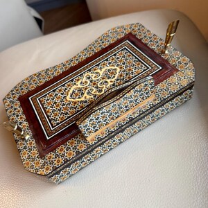 Pode incluir: Um conjunto de secret&aacute;ria de madeira ornamentado com um porta-canetas e uma pequena caixa. O conjunto apresenta um padr&atilde;o de mosaico detalhado em tons de castanho, dourado e preto. O porta-canetas tem uma caneta dourada. Projetado para organiza&ccedil;&atilde;o de secret&aacute;ria.