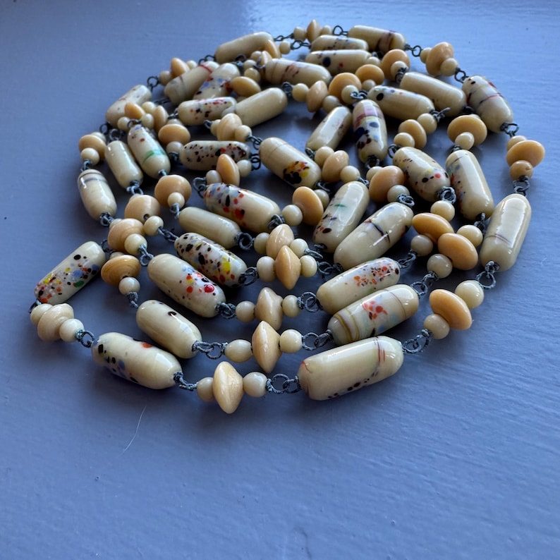 May include: A long necklace made of cream-coloured glass beads with colourful speckles. The beads are strung on a silver chain and interspersed with smaller, plain cream-coloured beads.