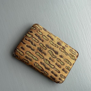 May include: A rectangular metal case featuring a textured fabric exterior with a repeating pattern of dark brown and light olive green abstract shapes. The case has rounded corners and a metallic gold-toned frame visible along the edges.