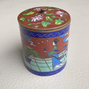 Puede incluir: Una caja cilíndrica de cloisonné antiguo con tapa. La caja es principalmente roja con una base azul y verde y una banda azul. La tapa presenta diseños florales rosas y blancos. El lateral de la caja representa a una mujer con vestimenta tradicional.