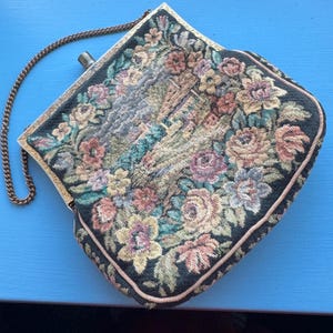 Puede incluir: Bolso de mano vintage de tapiz con un diseño floral en tonos rosa, amarillo y verde sobre un fondo negro. El bolso tiene un marco de metal dorado y una correa de cadena. Los bordes están ribeteados con un ribete rosa.