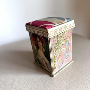 May include: A rectangular box with a padded lid featuring blue, beige, and magenta fabric panels. One side displays a vintage illustration of a person with red flowers in their hair, while the adjacent side features embroidered pink flowers and the text FOR AULD LANG SYNE.