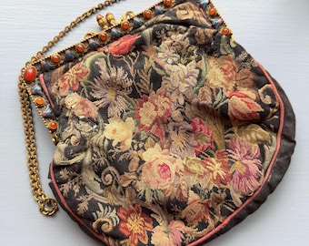 Könnte beinhalten: Eine Handtasche aus Gobelin mit dunklem Hintergrund und einem mehrfarbigen Blumenmuster. Die Tasche hat einen goldfarbenen Metallrahmen, der mit kleinen orangefarbenen Cabochon-Steinen verziert ist, und einen passenden Kettenhenkel.