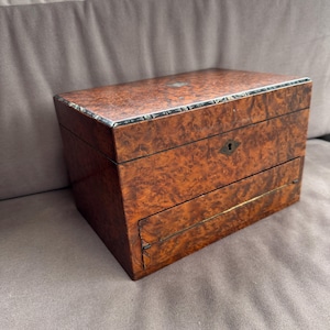 May include: A rectangular wooden box with a burl wood grain finish. The box has a small drawer and a keyhole. The edges are trimmed with a decorative border. The box is a warm brown colour.