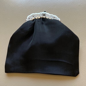 Puede incluir: Un bolso de noche de satén negro con un cierre de plata. El cierre tiene un diseño decorativo.