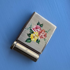 May include: A vintage gold-toned compact with a floral design. The compact features a textured surface and a hand-painted bouquet of pink, red, and yellow roses with green leaves. The lipstick holder is attached to the compact.