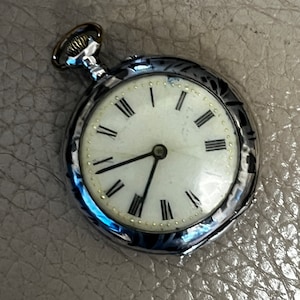 Puede incluir: Un reloj de bolsillo de plata con números romanos en la esfera y un diseño decorativo en la parte posterior.