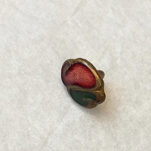May include: A small, antique button with a red and green enamel design. The button has a gold-colored metal frame and a textured surface. The red enamel is in the shape of a petal, and the green enamel is below it.