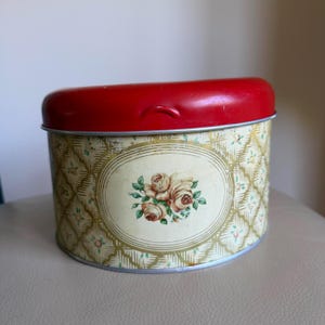 May include: Vintage metal tin with a red lid and a floral design. The oval center features a bouquet of roses. The tin's body has a cream background with a gold and green diamond pattern. A charming storage container.