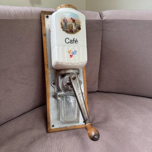 Puede incluir: Un antiguo molinillo de café de pared. Cuenta con un recipiente de cerámica blanca con tapón de corcho, una escena pastoral y la palabra "Café". El molinillo tiene un mecanismo de metal, un recipiente de vidrio y un mango de madera.