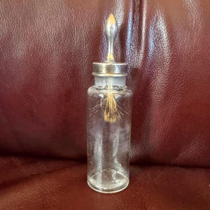 May include: A clear glass bottle with a silver-colored stopper and a brush inside. The bottle is cylindrical and has a silver metal cap. The brush is a light brown color. The bottle is likely for applying liquid.