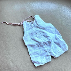 Könnte beinhalten: Ein weißer, ärmelloser Strampler mit Spitzenbesatz an den Beinöffnungen. Das Kleidungsstück hat eine rosa Schleife am Hals und ist aus leichtem Stoff gefertigt. Der Strampler ist für ein Baby oder Kleinkind konzipiert.