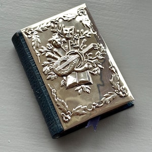 Pode incluir: Um pequeno livro prateado com uma capa em relevo apresentando um alaúde, máscaras teatrais e elementos florais. O livro tem uma lombada de couro azul e uma fita azul como marcador. O design é detalhado e decorativo.