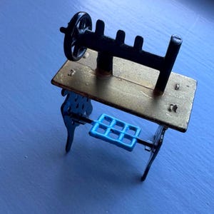 May include: A miniature sewing machine with a black wheel and frame, a brass-colored table, and a blue foot pedal. The legs of the table are black and have a decorative design. The sewing machine is a vintage-style collectible.