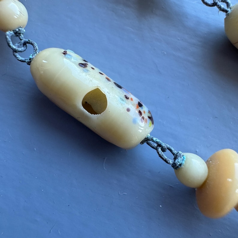 May include: A white glass bead necklace with a hole in the centre of each bead. The beads are speckled with brown, orange, and blue flecks.