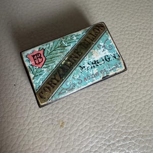 May include: A vintage matchbox with a blue and green design. The text on the box reads "Cory Line Billon" and "Dess Marjgni" with a small crest in the upper left corner.