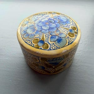 May include: A circular trinket box with a cream-coloured base and lid. The lid is adorned with a central floral design in shades of blue and green. The sides of the box feature a repeating floral pattern, giving it a vintage appearance.