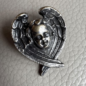 Puede incluir: Un broche de ángel plateado de estilo antiguo y detallado. El broche presenta una cara de querubín con alas detalladas. El metal tiene un aspecto ligeramente deslustrado, lo que aumenta su encanto vintage. El broche probablemente está hecho de una aleación de metal.