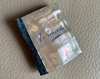May include: A small, rectangular mother-of-pearl item with a blue fabric spine. The surface has iridescent qualities and features the cursive word "Couville" in blue ink. The item appears to be a small book or decorative piece.