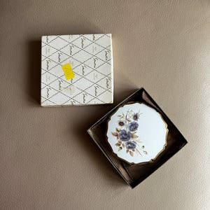 Pode incluir: Um espelho compacto Stratton vintage com um design floral numa caixa. O compacto tem um fundo branco com flores azuis e acabamento dourado. A caixa é branca com um padrão de diamante e a palavra "Stratton" repetida.