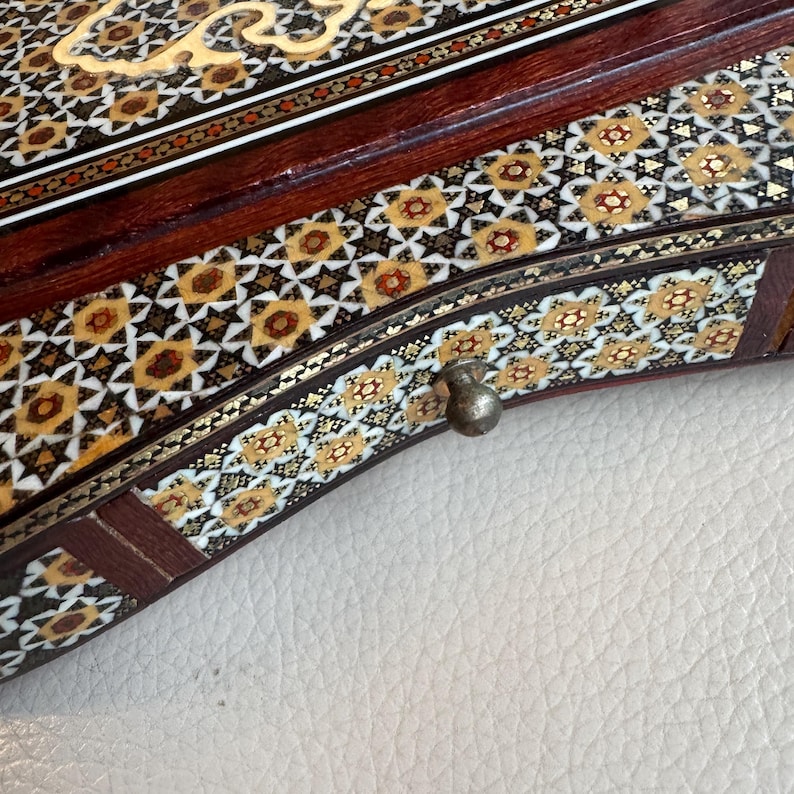 Pode incluir: Vista de perto de uma caixa de madeira com intrincados padr&otilde;es de mosaico embutidos. A caixa apresenta madeira castanha escura com desenhos geom&eacute;tricos em dourado, branco e castanho. Uma pequena pega de metal ornamentada &eacute; vis&iacute;vel na borda curva.