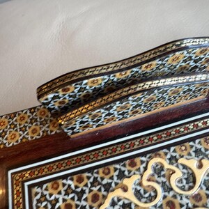 Pode incluir: Close-up de uma caixa de madeira decorativa com intrincados padr&otilde;es embutidos. A caixa apresenta uma base marrom escura com desenhos geom&eacute;tricos em ouro, branco e vermelho. A al&ccedil;a curva e as bordas s&atilde;o adornadas com padr&otilde;es detalhados semelhantes.