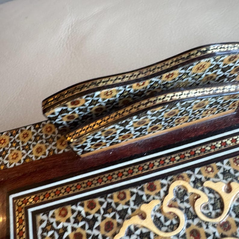 Pode incluir: Close-up de uma caixa de madeira decorativa com intrincados padr&otilde;es embutidos. A caixa apresenta uma base marrom escura com desenhos geom&eacute;tricos em ouro, branco e vermelho. A al&ccedil;a curva e as bordas s&atilde;o adornadas com padr&otilde;es detalhados semelhantes.