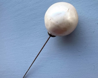 May include: A vintage hat pin with a round, off-white pearlized head and a long, slender, dark metal pin. The pin is set against a light blue background.