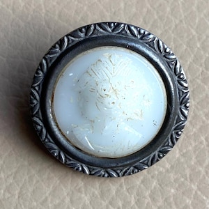 Peut inclure: Un bouton en cam&eacute;e blanc avec un cadre en m&eacute;tal argent&eacute;. Le cam&eacute;e pr&eacute;sente un motif en relief d'un visage de femme.