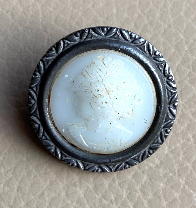 Peut inclure: Un bouton en cam&eacute;e blanc avec un cadre en m&eacute;tal argent&eacute;. Le cam&eacute;e pr&eacute;sente un motif en relief d'un visage de femme.