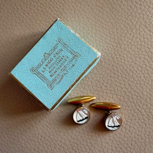 Puede incluir: Gemelos dorados con diseño de velero, junto a una caja azul claro con ribetes dorados. La caja lleva la inscripción "AS WIGG & SON JEWELLERS HIGH STREET NEWMARKET."