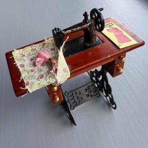 May include: Miniature sewing machine with a dark wood table and black metal base. The machine has a black wheel and red accents. A small piece of fabric with pink thread and a floral pattern sits on the table. A small card with a pink dress illustration is also on the table.
