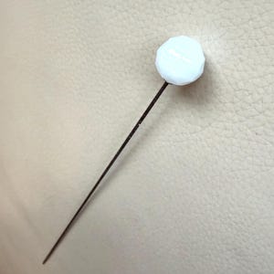 Puede incluir: Un alfiler de sombrero con una parte superior blanca y un eje metálico oscuro y delgado. La cabeza blanca tiene un diseño geométrico facetado. El alfiler está diseñado para asegurar sombreros u otros accesorios.