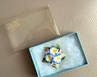 May include: A Fenton China English Bone China floral brooch in a light blue box with a clear plastic cover. The brooch features blue, yellow, and pink flowers with green leaves. The box is lined with white cotton.