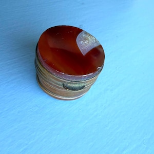 May include: A small, round metal container with a polished, deep reddish-orange stone lid. The stone features a translucent, crystalline inclusion on one side. The base of the container is made of a muted, weathered brass-toned metal with a simple latch.