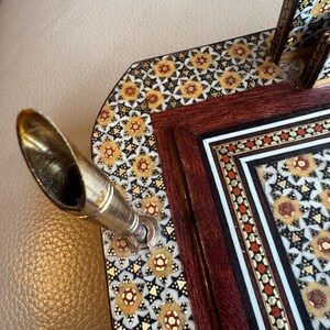 Pode incluir: Close-up de um porta-canetas decorativo com uma caneta prateada e um intrincado design de mosaico. O porta-canetas apresenta uma moldura de madeira escura com padr&otilde;es incrustados de ouro, vermelho e branco. O design do mosaico &eacute; um padr&atilde;o floral repetitivo.