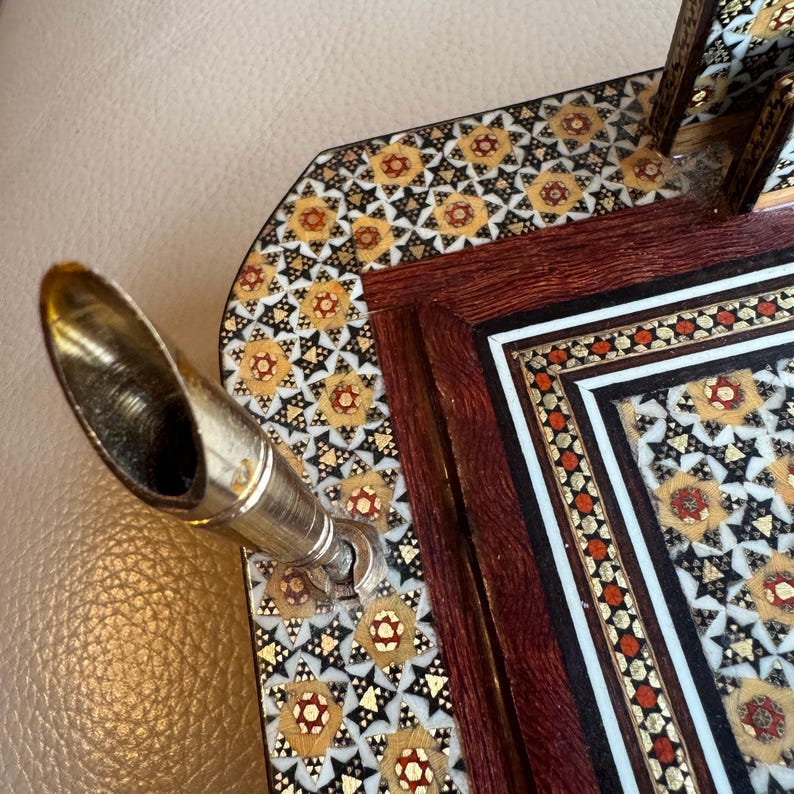 Pode incluir: Close-up de um porta-canetas decorativo com uma caneta prateada e um intrincado design de mosaico. O porta-canetas apresenta uma moldura de madeira escura com padr&otilde;es incrustados de ouro, vermelho e branco. O design do mosaico &eacute; um padr&atilde;o floral repetitivo.