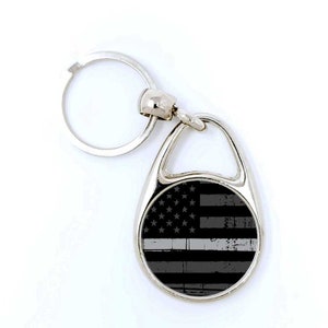 May include: A silver metal keychain with a black and white thin blue line American flag design. The keychain has a split ring and a hook.