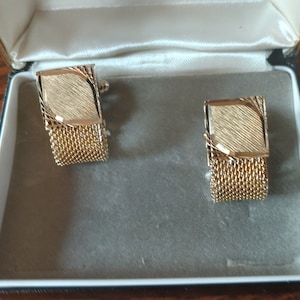 May include: A pair of gold cufflinks with a mesh design and a textured gold top. The cufflinks are in a black and white box.