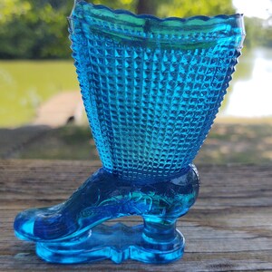May include: A blue glass boot-shaped vase with a textured diamond pattern. The vase is made of clear glass with a blue tint.